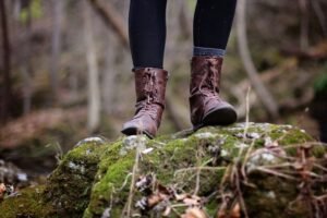 Close-up of brown boots hiking on mossy rocks in a scenic forest setting.
